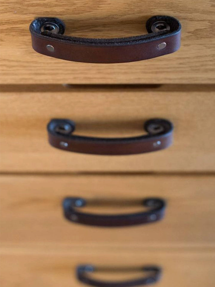 Brown leather drawer pulls on light-colored wooden furniture.  Curved handles, metal fasteners.