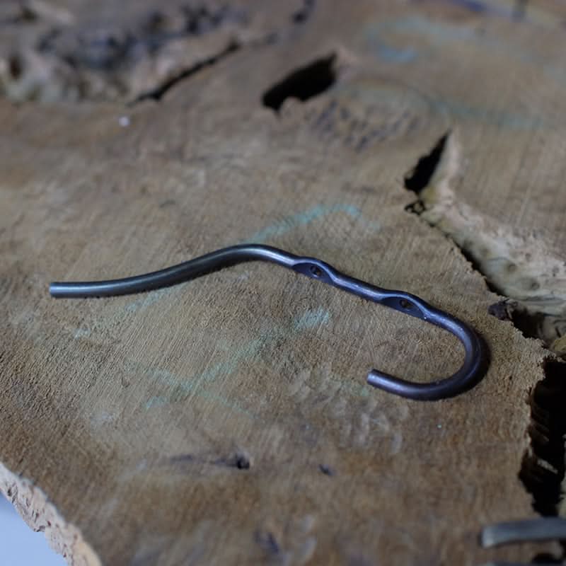 Rustic wall hook, hand-forged iron, dark metal finish on wood grain background.