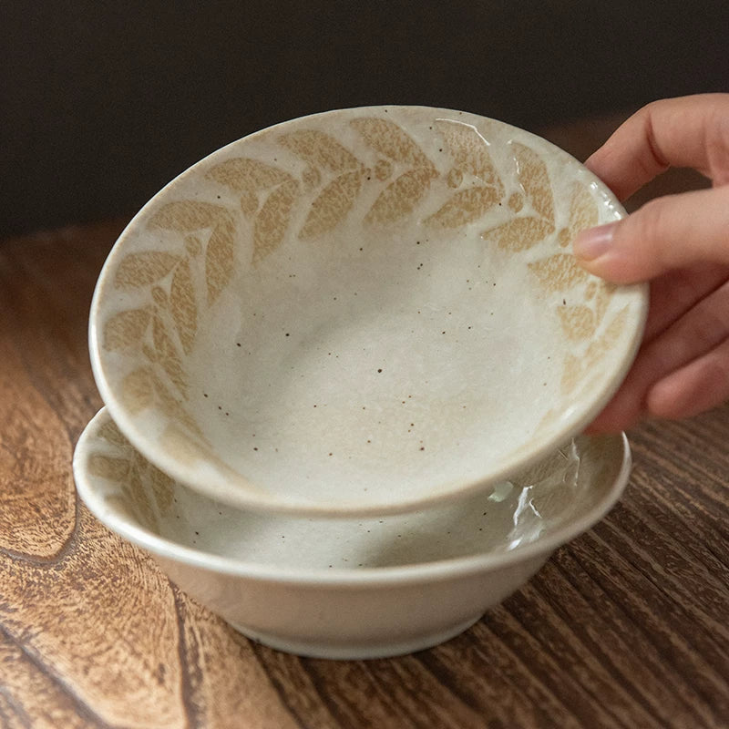 Close-up of two cream ceramic bowls with a beige leaf pattern being stacked by hand.