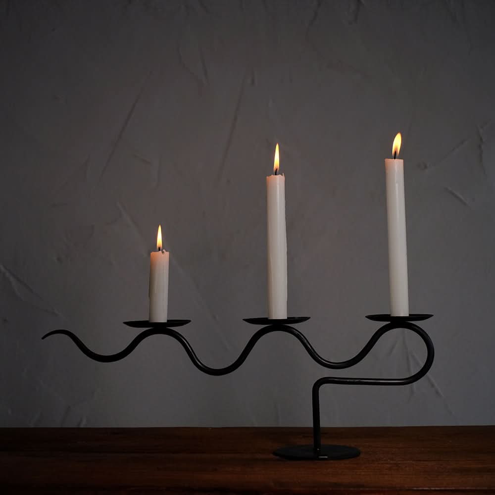 Black metal Three-Candle Holder with wavy design and lit white candles on wooden surface.