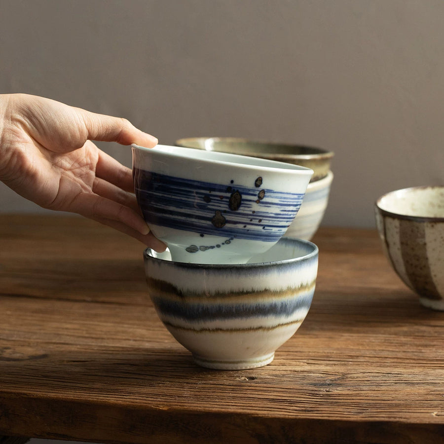 Hands stacking two rustic ceramic rice bowls with blue brushstroke patterns on a wooden table.
