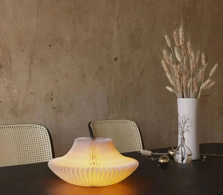 Origami bell-shaped paper bedside lamp glows warmly on a table, beside wheat in a vase.