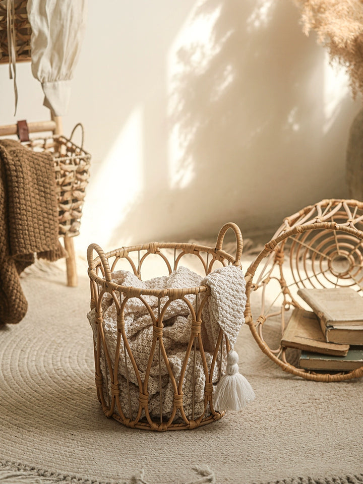 Round woven rattan storage basket, light beige color, with handles and a cream-colored knitted throw inside.  Stylish decorative basket on a beige rug.