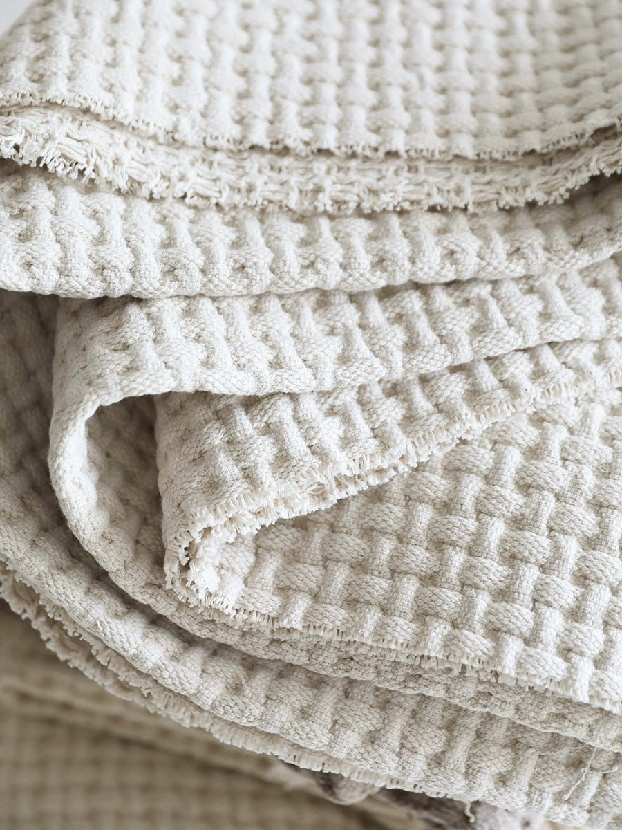Beige woven cotton throw blanket with textured weave and fringed edges. Cozy home decor.