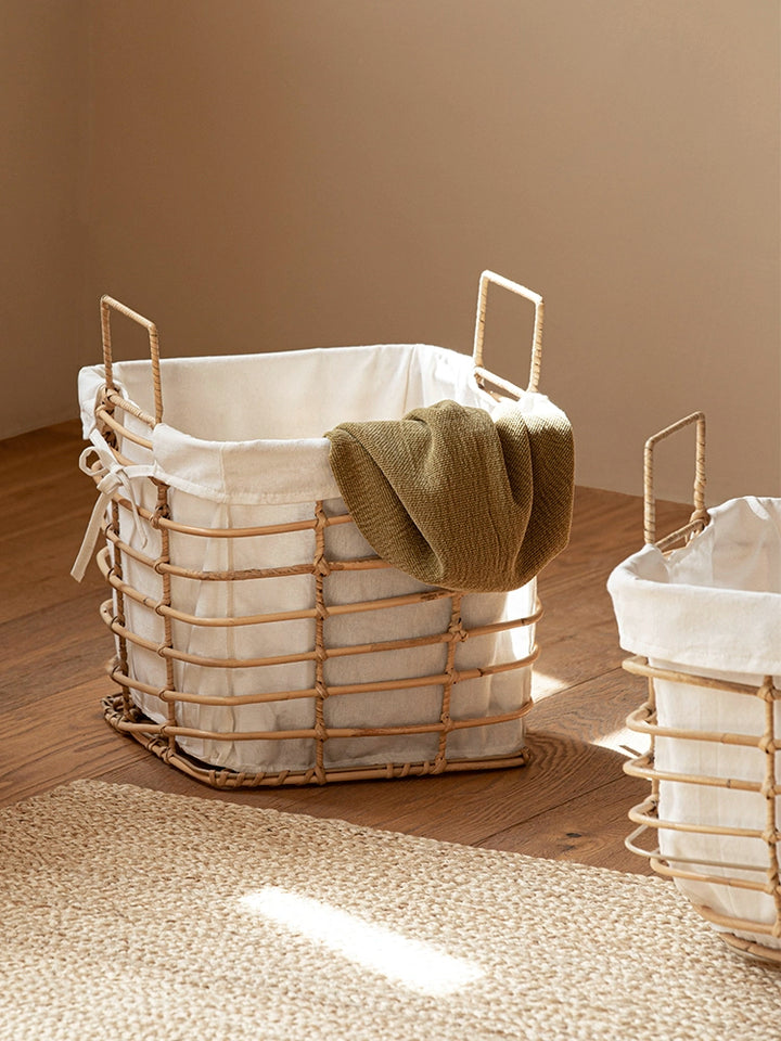 Square woven rattan laundry basket with cream linen liner.  Beige basket, light-colored rug, and a olive-toned garment visible inside.