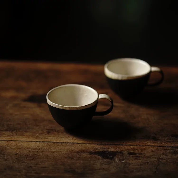 Two artisanal black and cream ceramic teacups arranged on a dark rustic wooden table, soft dramatic lighting.