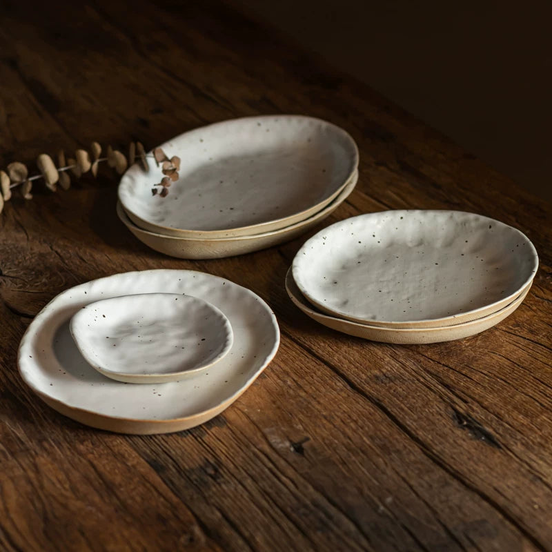 Set of speckled, cream-colored ceramic dishes and bowls stacked on a rustic wooden table.
