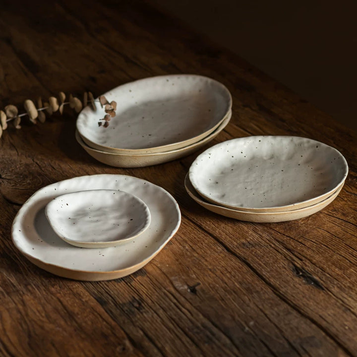 Set of speckled, cream-colored ceramic dishes and bowls stacked on a rustic wooden table.
