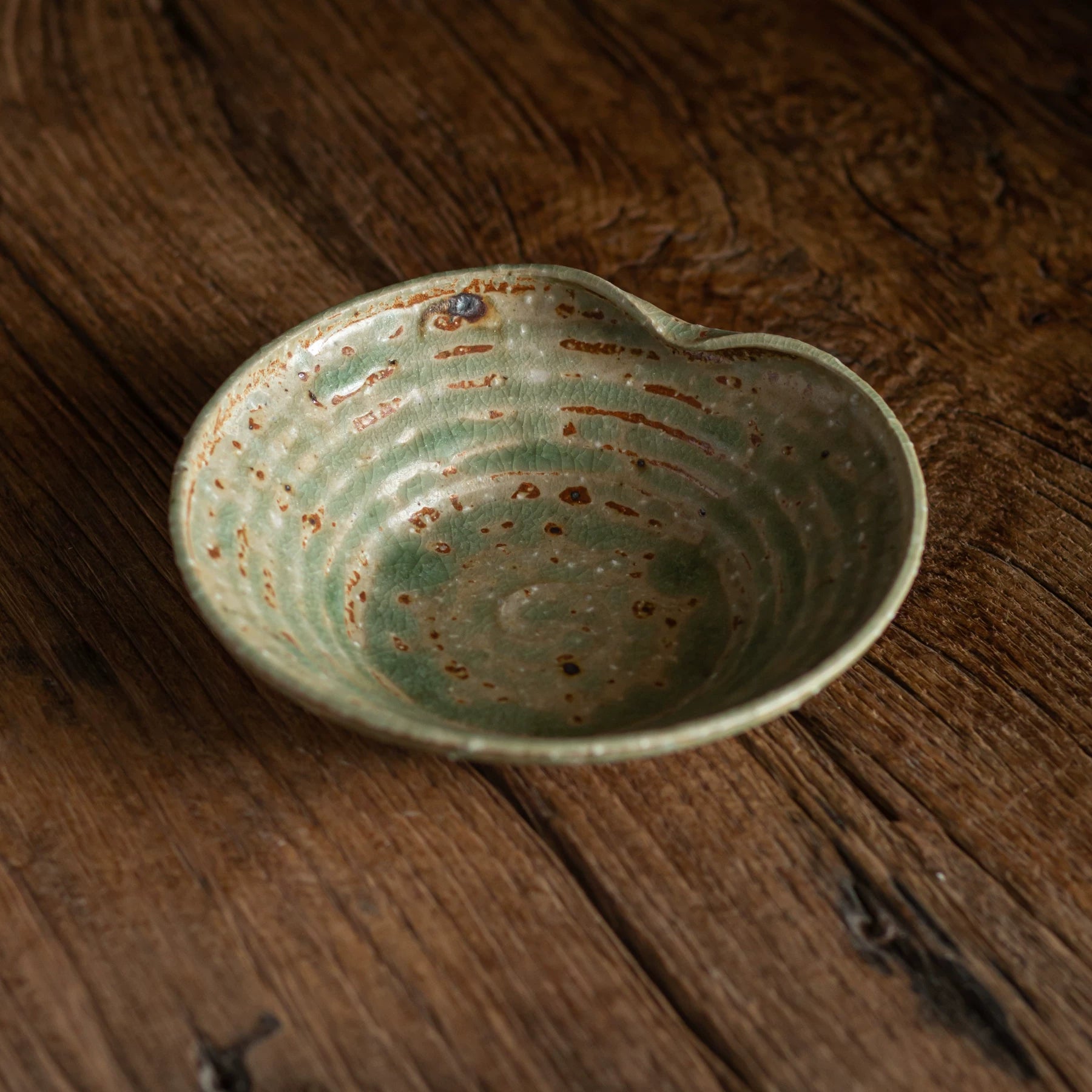 Close-up of a rustic, heart-shaped ceramic bowl with green and brown reactive glaze on a wooden table.