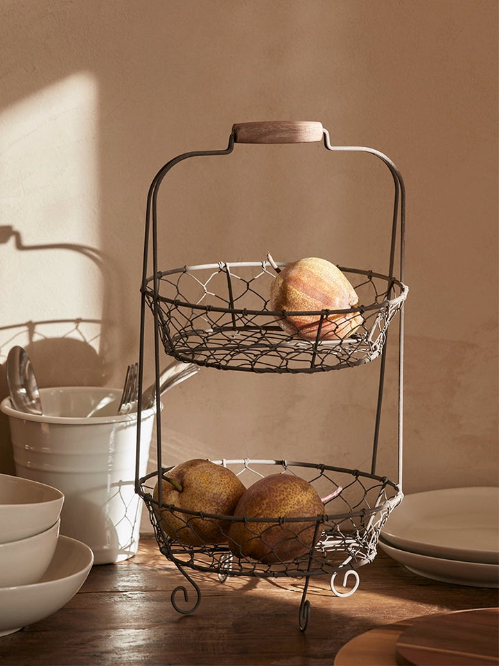 Two-tiered wire fruit basket stand, rustic style.  Dark gray wire, wooden handle, and oval wire baskets hold pears.  Kitchen decor.