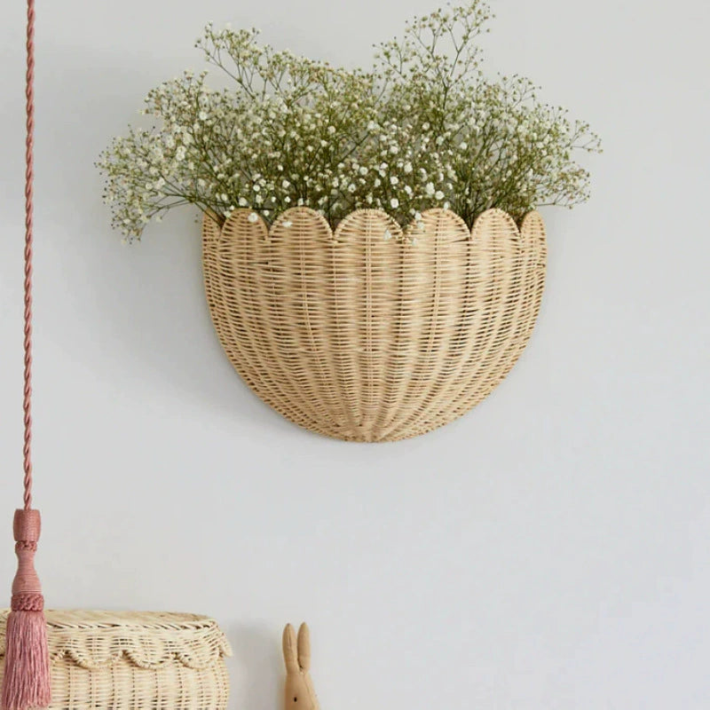 Beige scalloped wicker wall basket, holding small white flowers.  Natural woven wicker.  Circular shape.  Mounted on a wall.