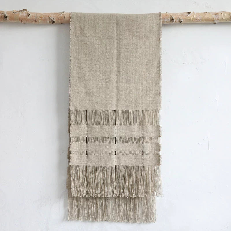 Beige linen table runner with tassels, rustic woven decor hanging on a branch against a white wall.