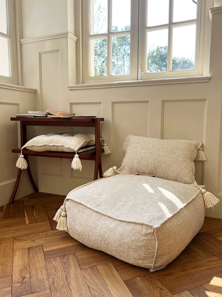 Beige embroidered ottoman pouf with tassels on wooden floor, decorative floor cushion for living room