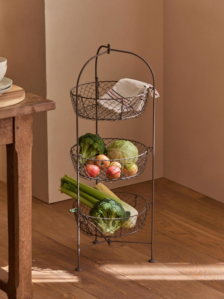 Rustic 3-tier wire fruit basket stand with metal wire baskets holding produce.  Dark brown/bronze finish.  Wooden floor.