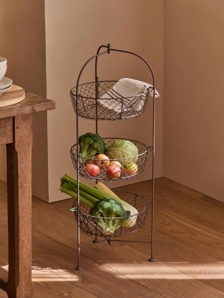 Rustic 3-tier wire fruit basket stand with metal wire baskets holding produce.  Dark brown/bronze finish.  Wooden floor.