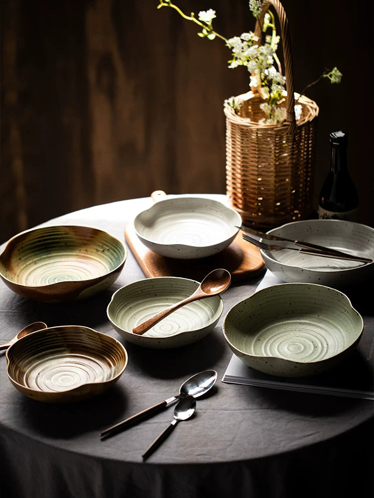 Collection of rustic, irregular ceramic dishes in brown, cream, and green on a draped table setting.