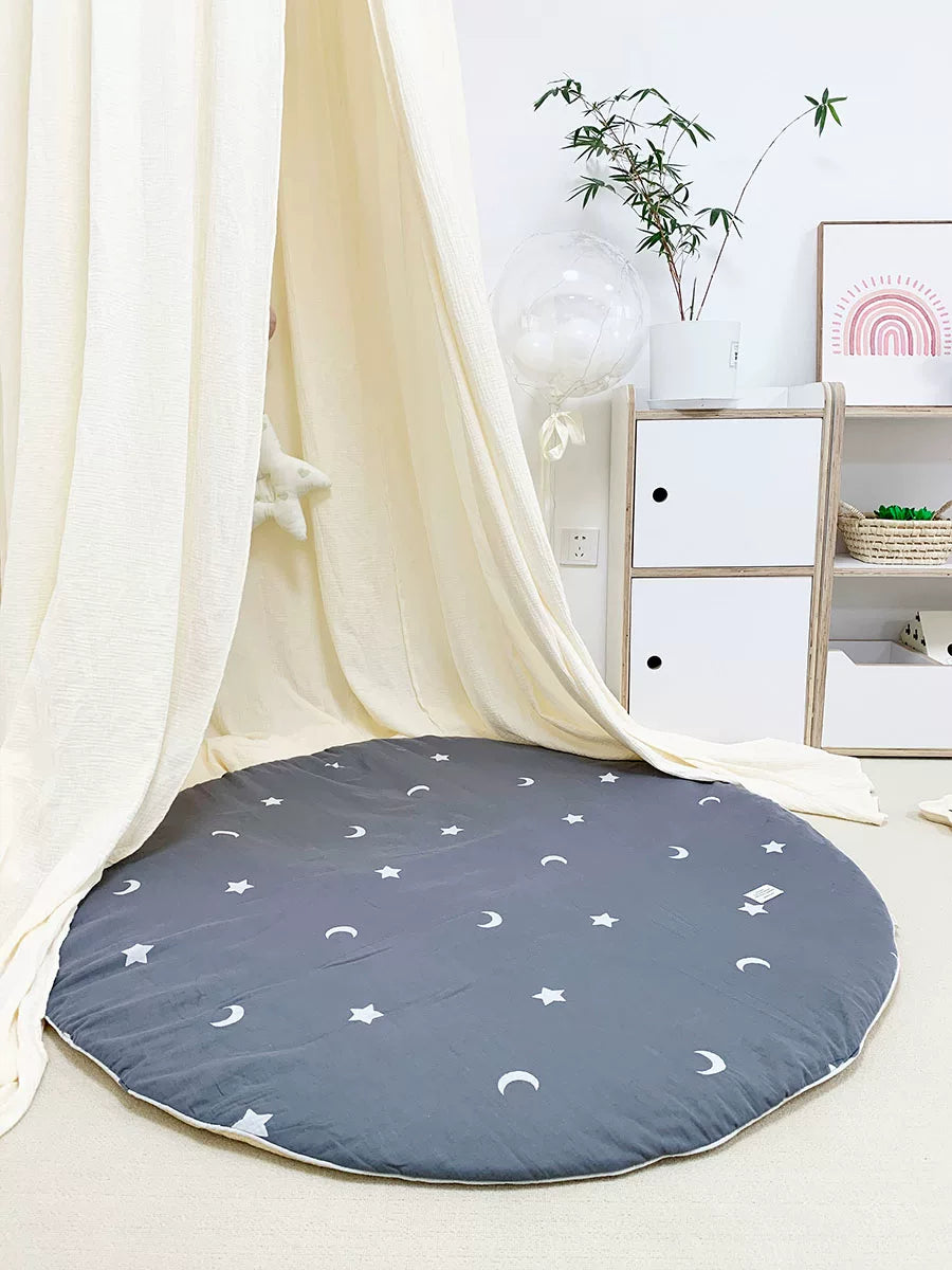 Round dark grey play mat with white stars and moons underneath a sheer white canopy.