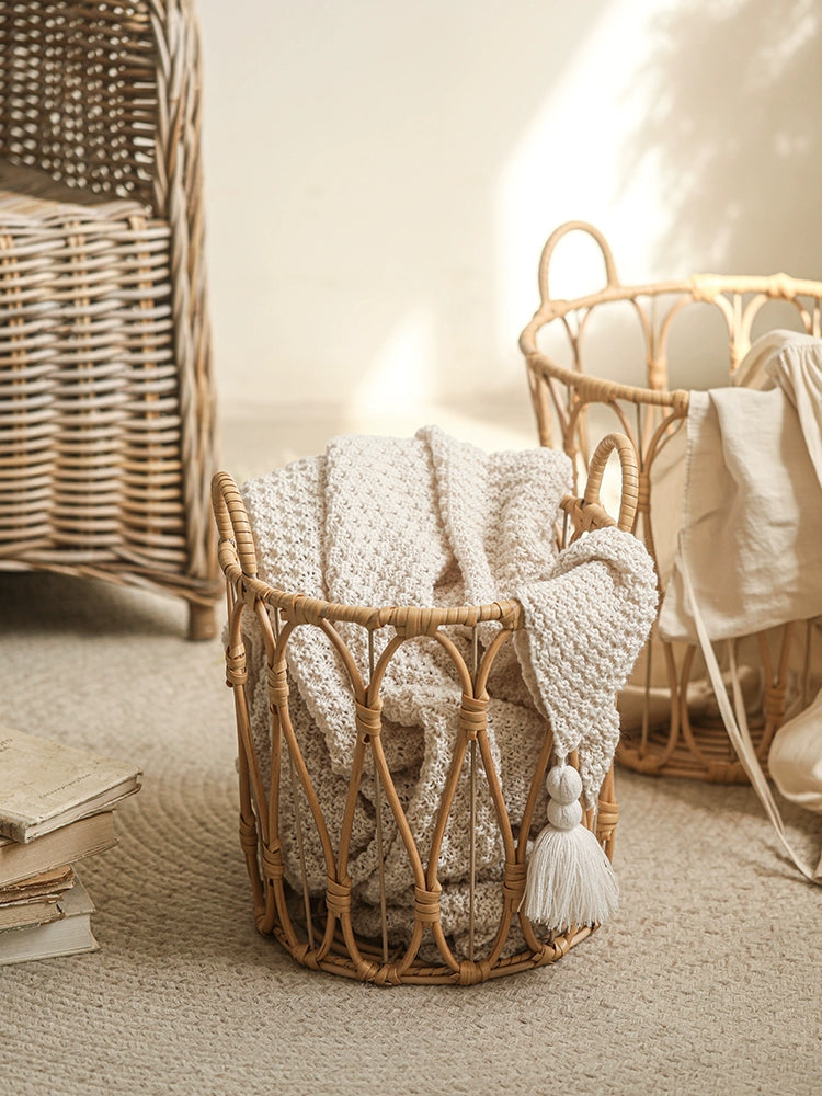 Light beige woven rattan storage basket with cream-colored knitted blanket inside.  Round basket with metal accents and handles.  Home decor.
