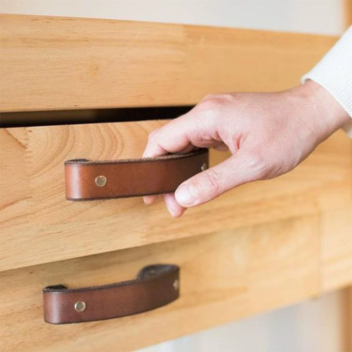 Brown leather drawer pulls on light-colored wooden furniture.  Curved handles with metal rivets.