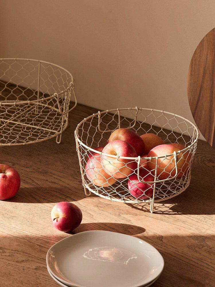 Beige wire dish basket with handles, holding apples on a wooden surface.