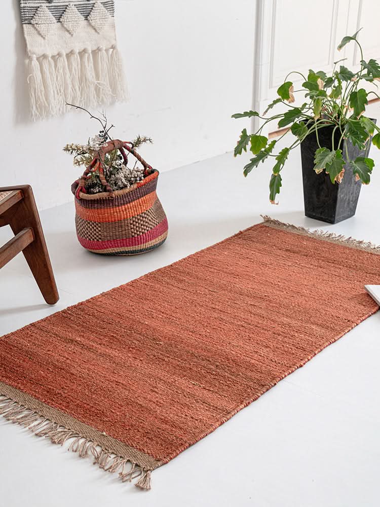 Handwoven Jute Rug - Natural Rustic, terracotta color, fringed edges, with woven basket and plant.