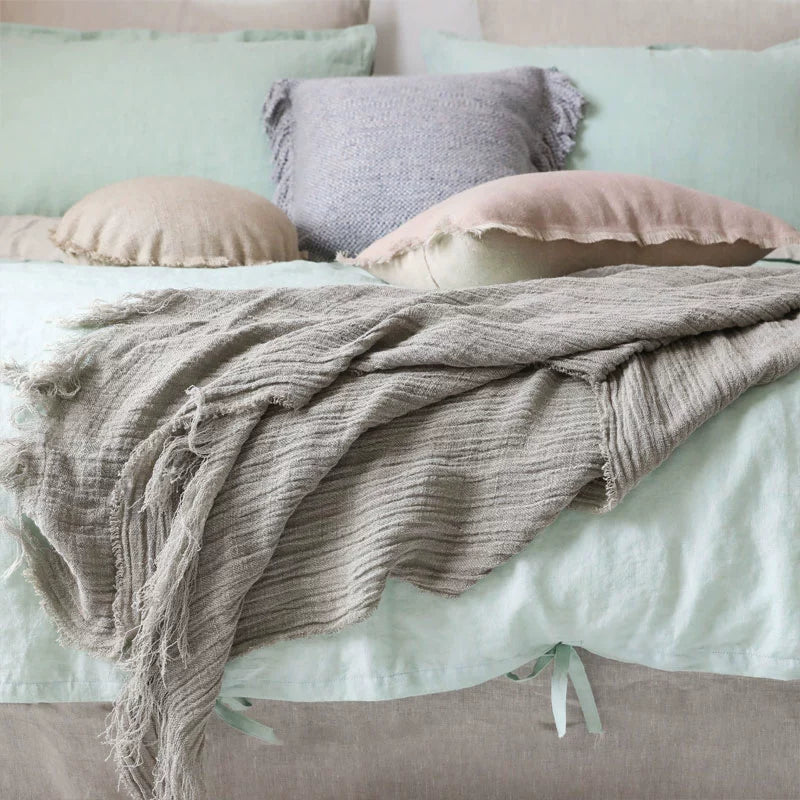 Light gray linen throw blanket with frayed fringes, draped over a mint green bed.  Soft textured linen.