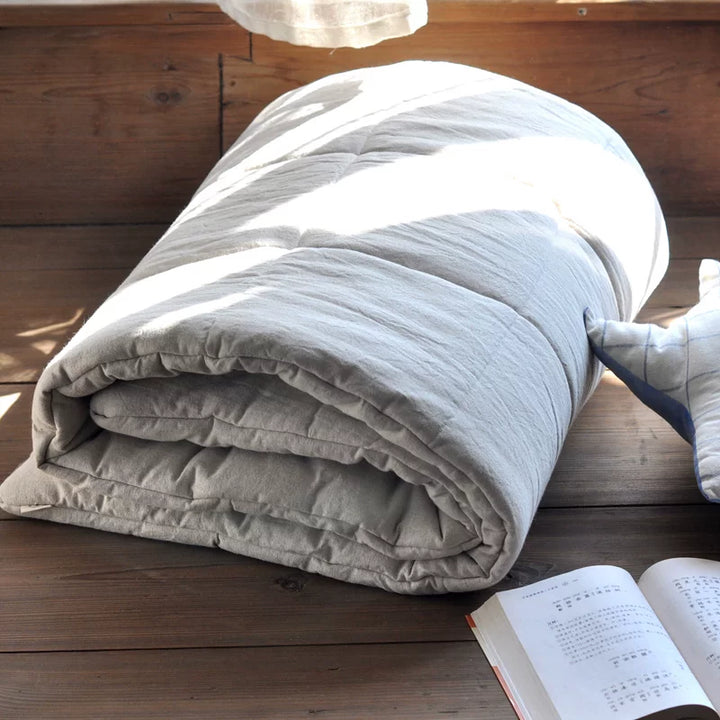 A light beige/off-white linen comforter or futon casually rolled up on a wooden floor with an open book.