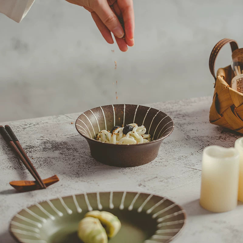 Close-up of a hand sprinkling seasoning over food in a brown ceramic bowl with white stripes on a light table.