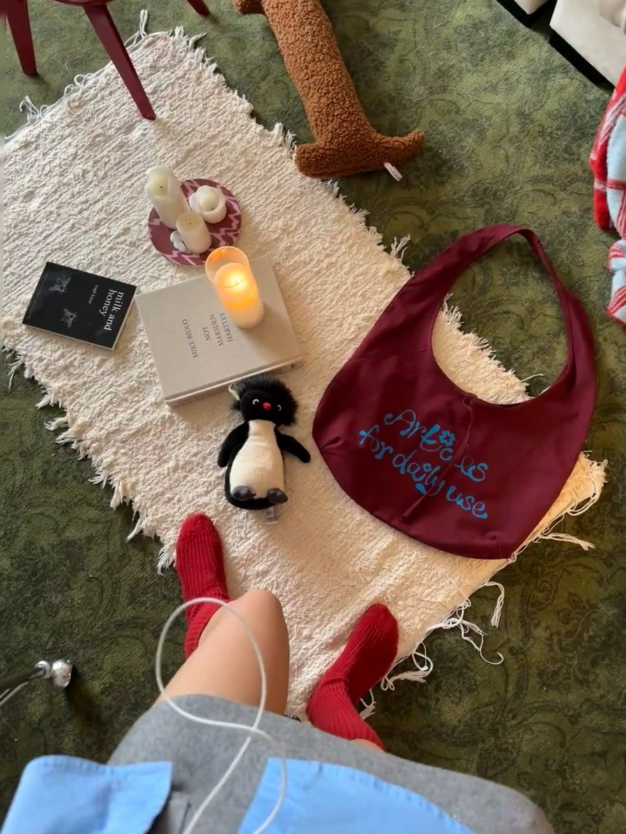 Beige knitted throw blanket, boho style, on a cream rug with candles, books, and a stuffed penguin.  Red socks and a person's legs are visible.  Dark maroon bag with text.