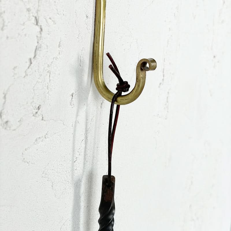 Gold J-shaped wall hook made of brass on a textured white wall with a dark cord detail.