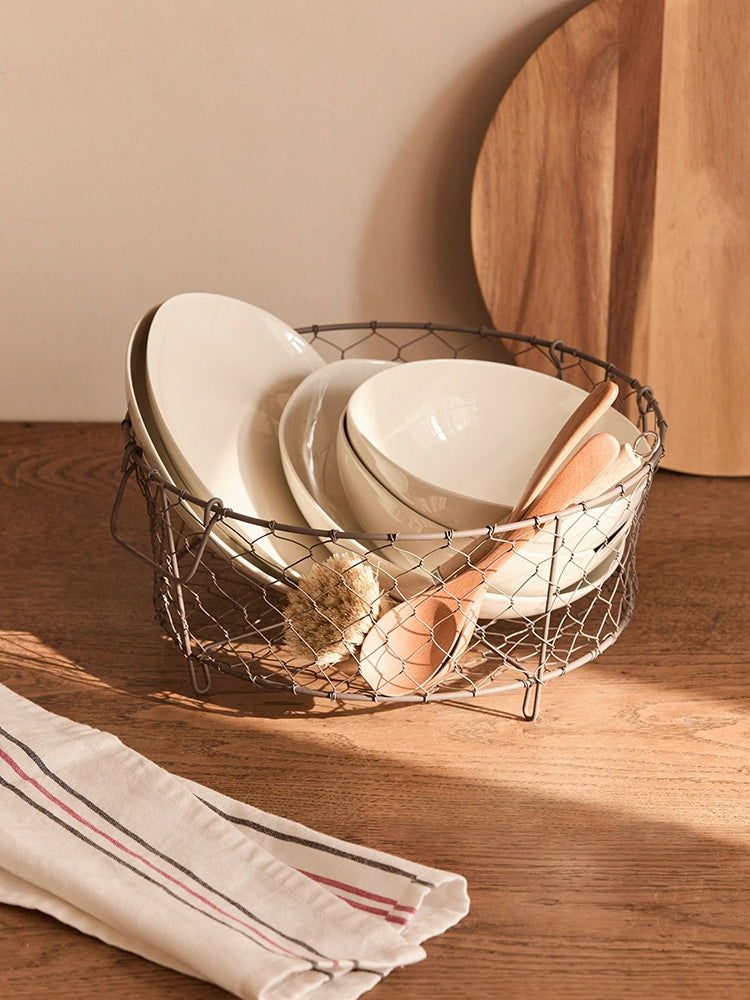 Beige wire dish basket with handles holding white dishes, wood spoons, and a scrub brush on a wooden surface.