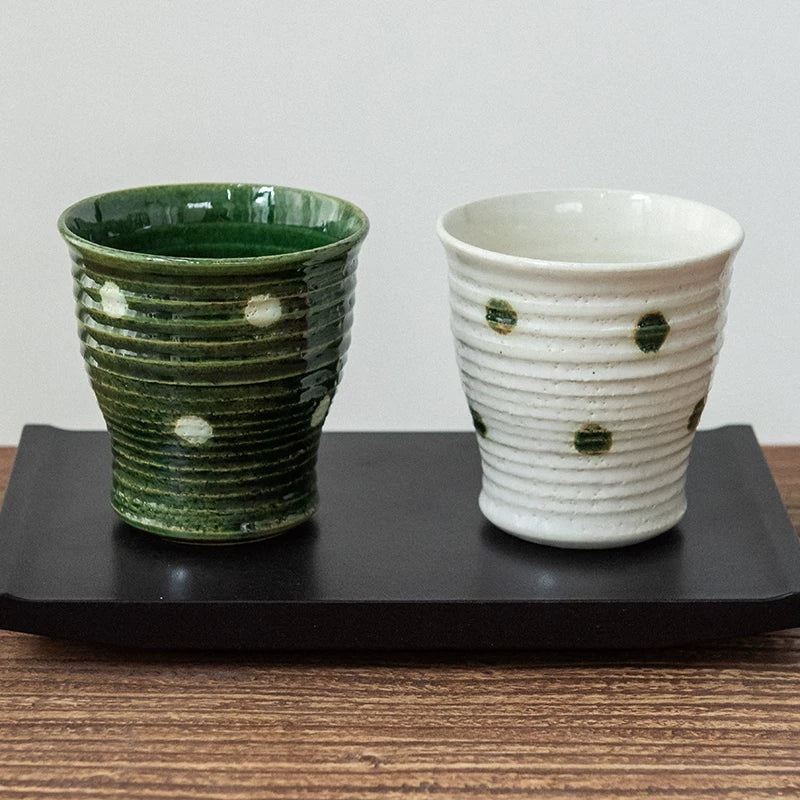 Two ceramic Yunomi tea cups, one green and one white, with dark green polka dots, sitting on a black tray.