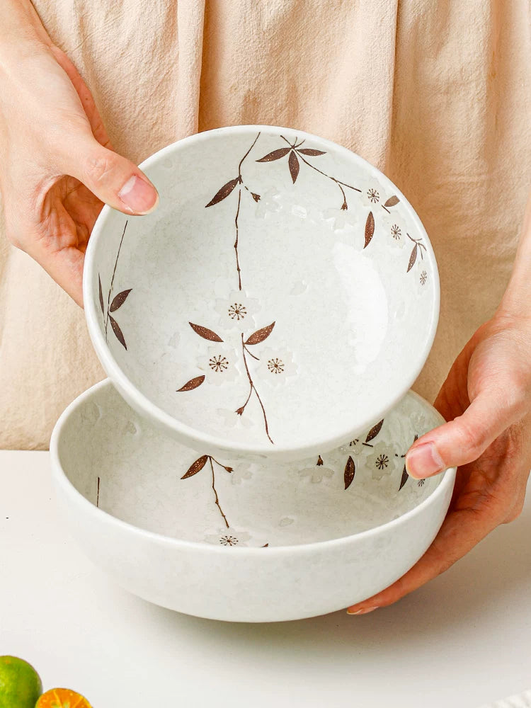 Hands holding two white ceramic bowls with delicate brown cherry blossom pattern, product overview.