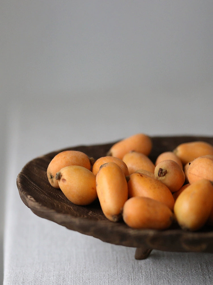 Fresh loquats on a dark wooden platter with carved legs.  A light-colored linen tablecloth is beneath the platter.