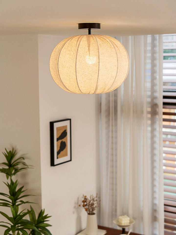 Beige linen round ceiling lamp with pleated design.  Warm light, modern style.  Mounted in a room with plants, art, and sheer curtains.