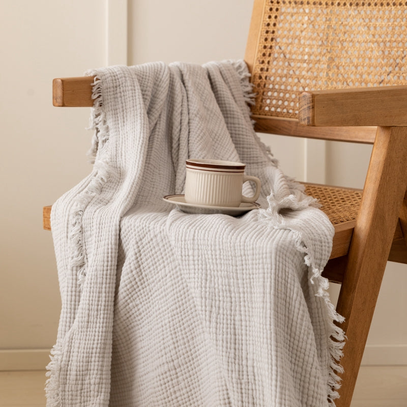 Light gray waffle weave throw blanket draped over wooden chair.  Frayed edges.  Small cup on saucer.