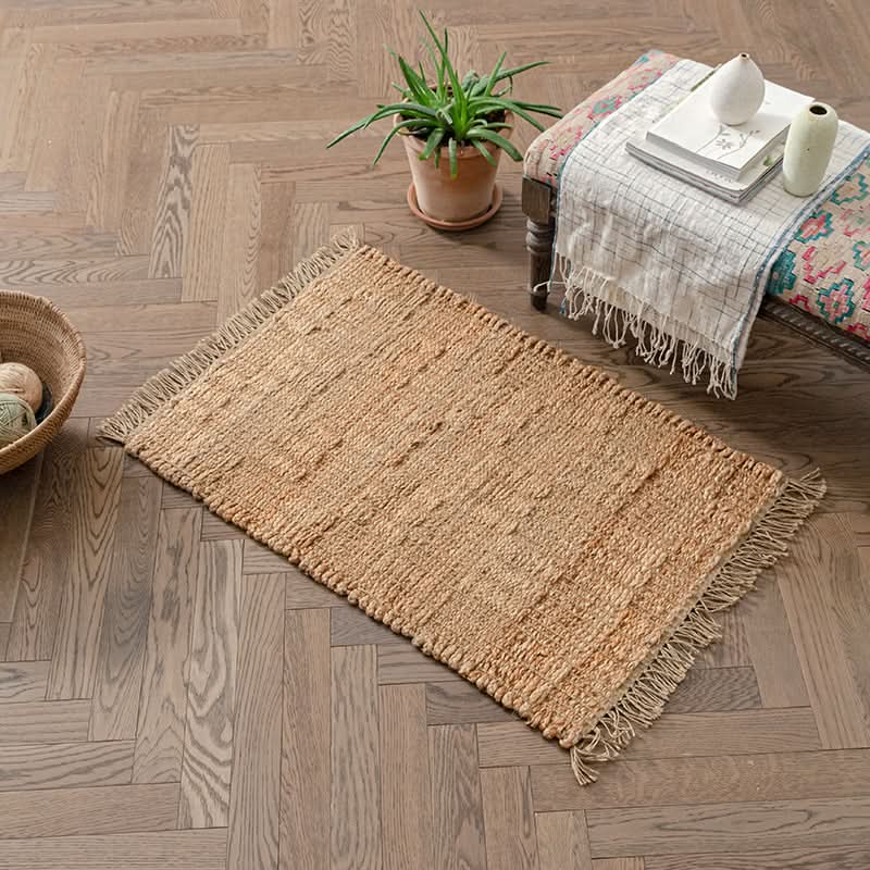 Natural woven area rug with fringe on wood floor, near plant and bench.