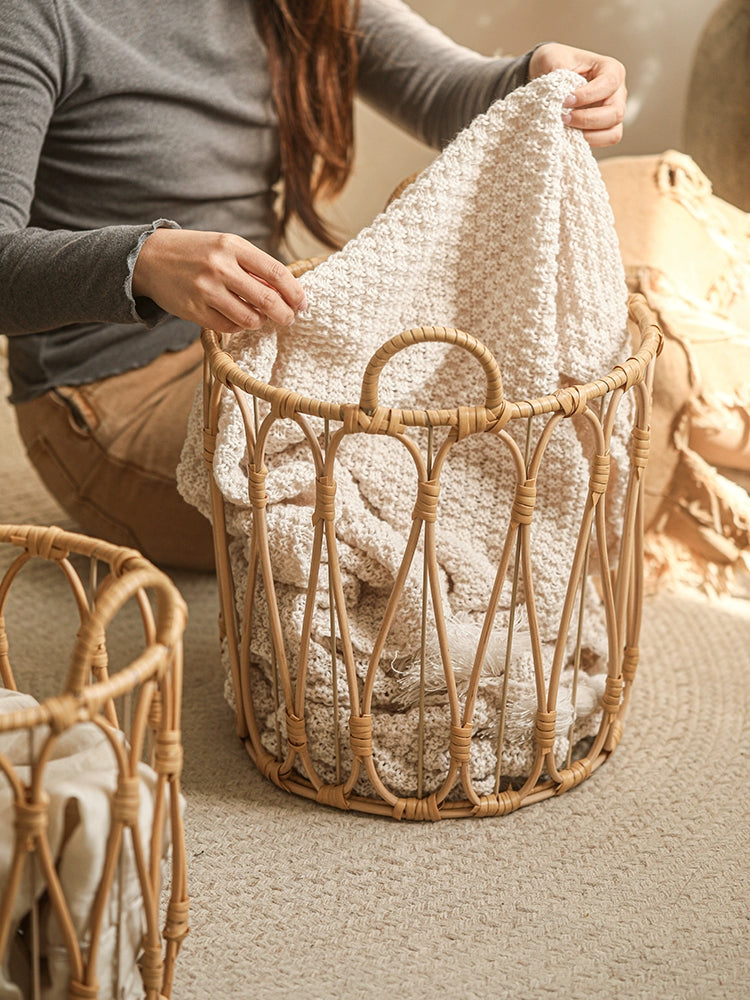 Round woven rattan storage basket, light beige color, with handles, filled with a cream-colored knitted blanket.  Handcrafted, decorative.