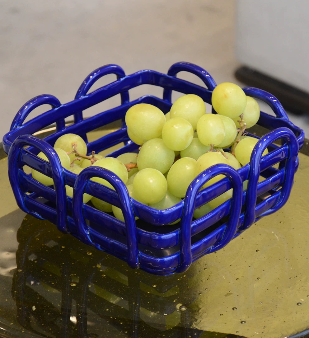 Blue ceramic fruit basket with woven design.  Square shape, vibrant blue color, holding green grapes.  Stylish serving dish.