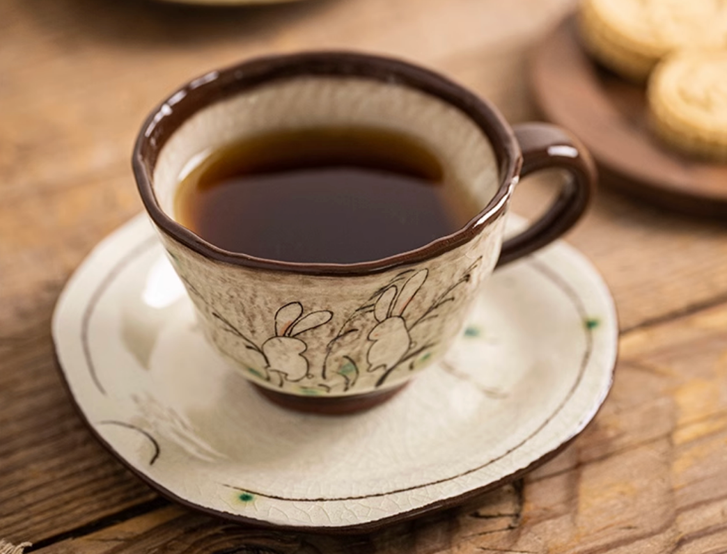 Handmade ceramic rabbit mug with coaster, beige with brown trim, filled with tea.