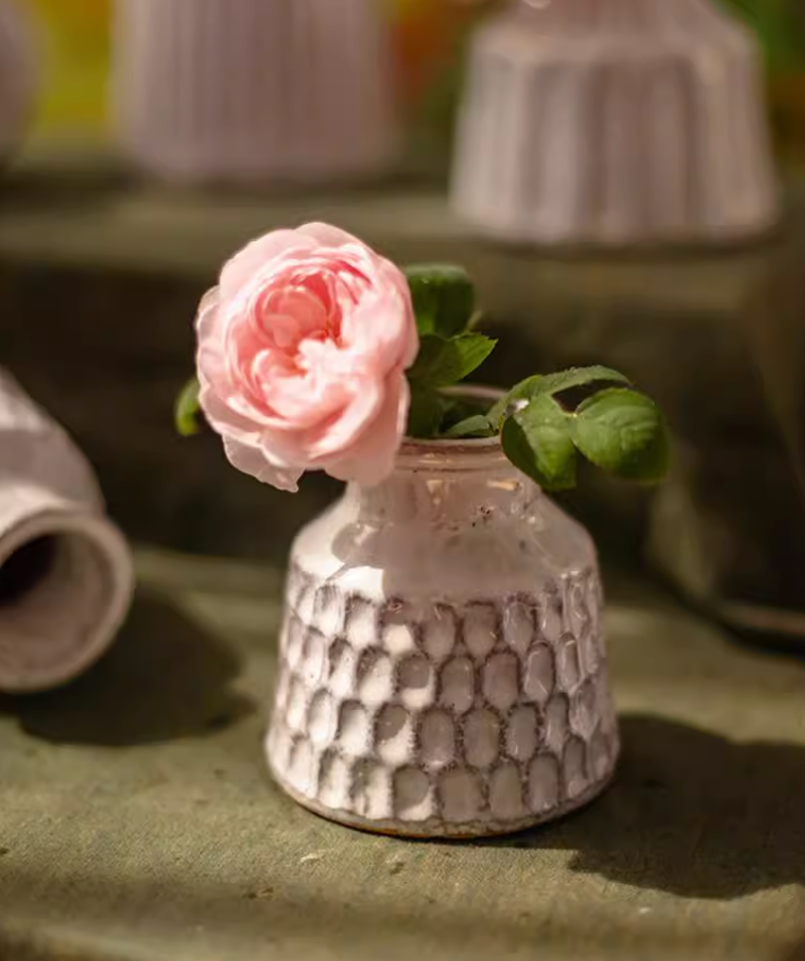White ceramic small vase with honeycomb texture holding a pink rose on a textured green surface.