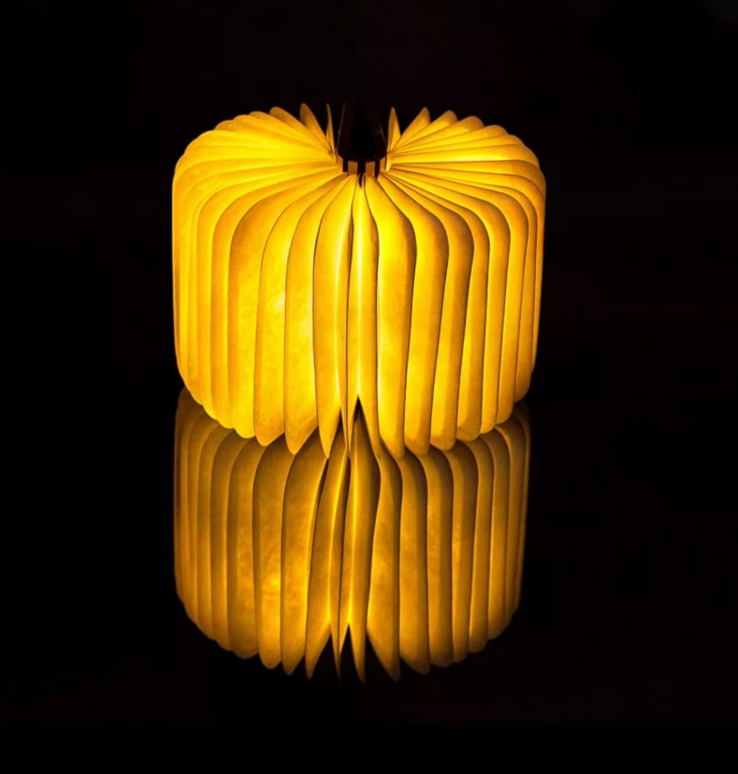 Warm yellow light glows from this Customizable Book Paper Table Lamp, reflecting on a dark surface.