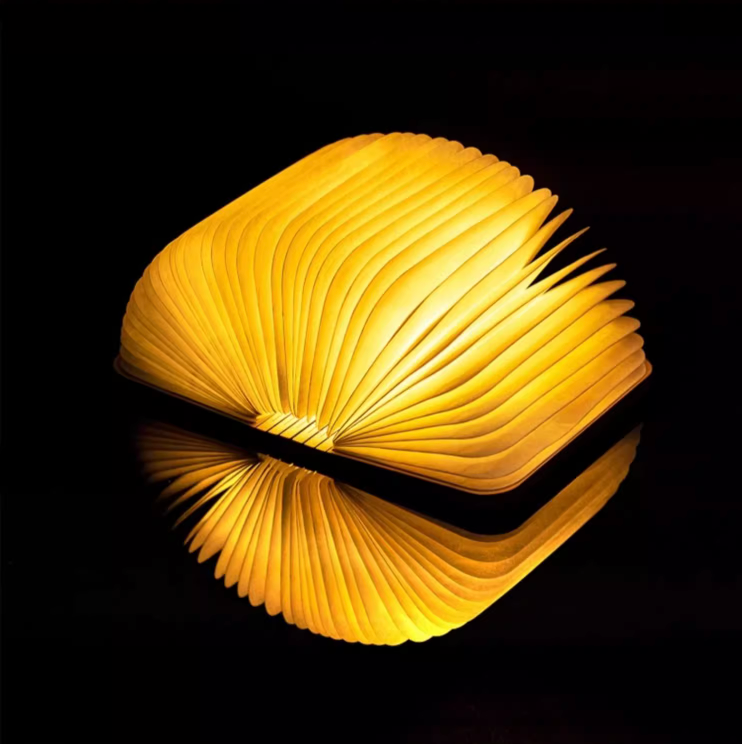 Illuminated Customizable Book Paper Table Lamp glows warm yellow, reflecting on dark surface.