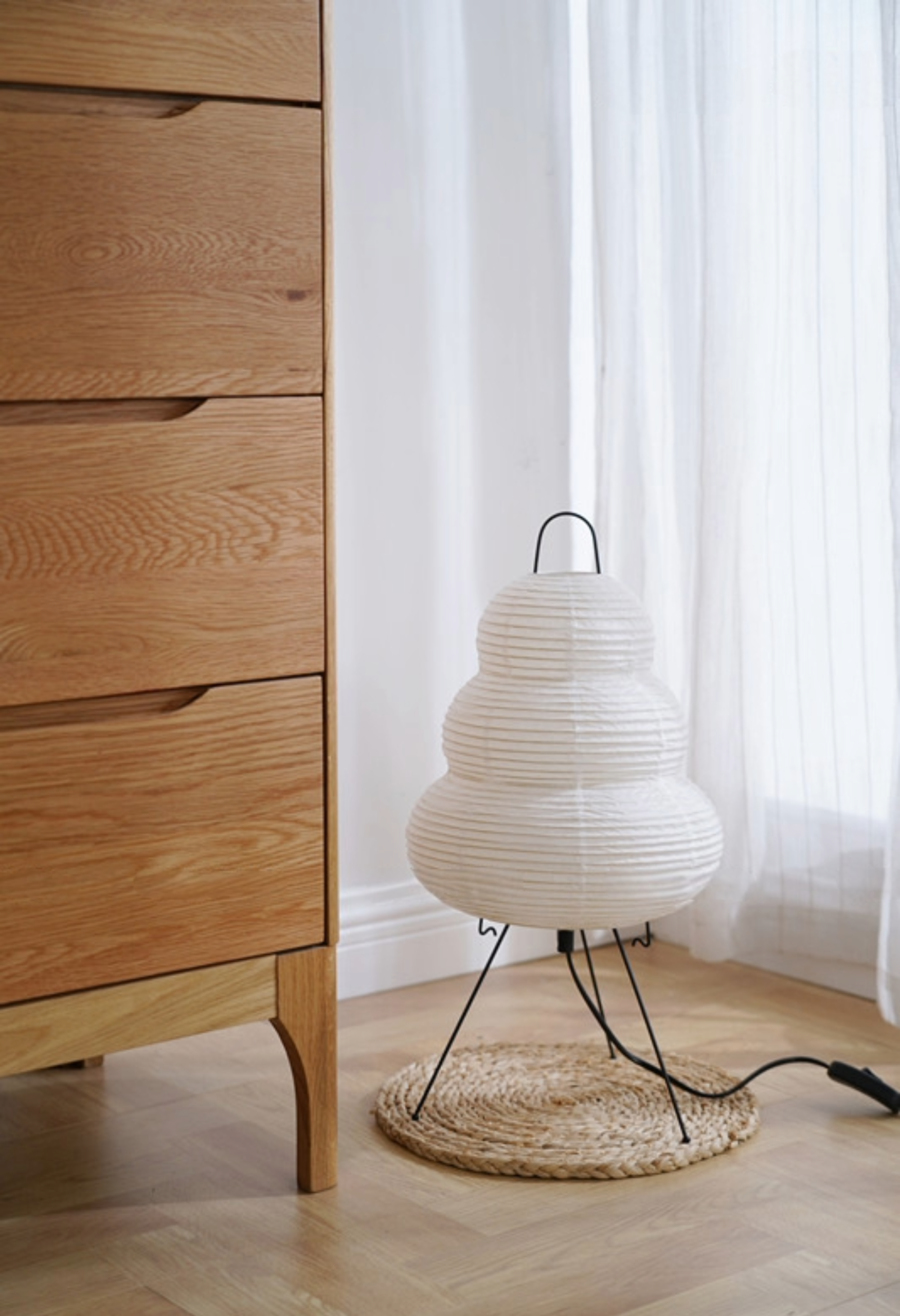 White tiered paper floor lamp with black legs on a woven mat next to a wooden dresser.