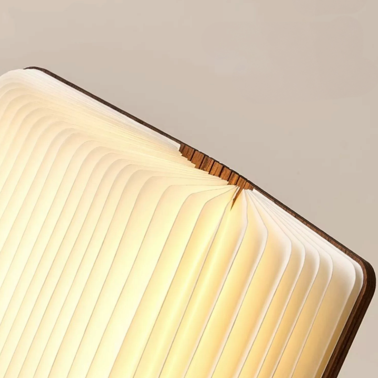 Warm white light emanates from a closed folding portable paper lamp with wooden covers.