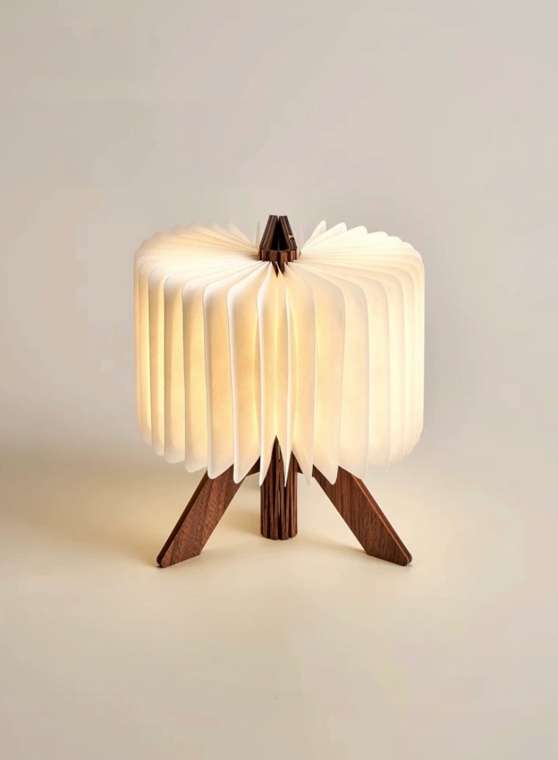Illuminated white paper folding portable lamp with a wooden tripod base on a neutral background.