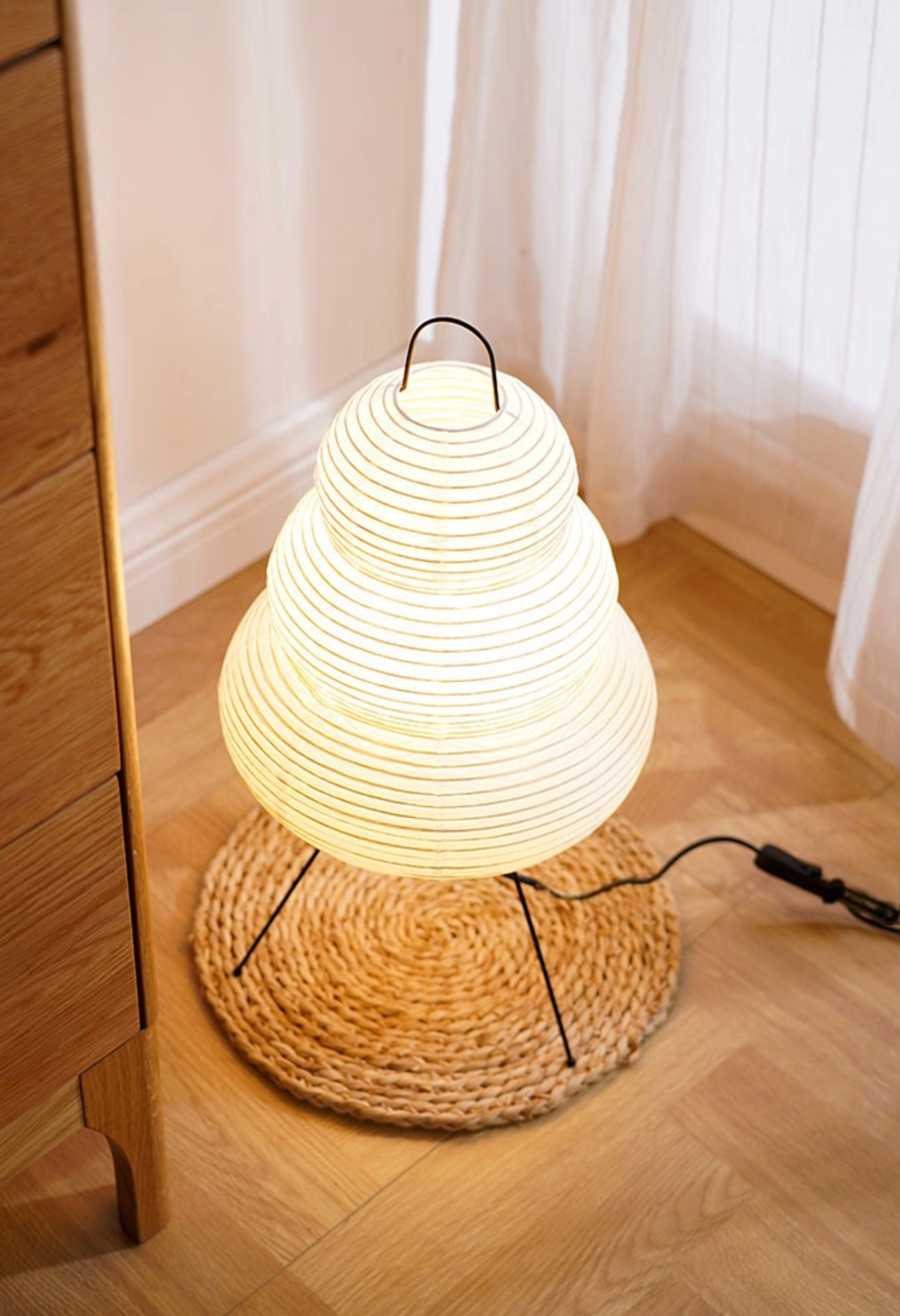 Warm light glows through this tiered Floor Paper Lamp on a woven mat next to wood furniture.