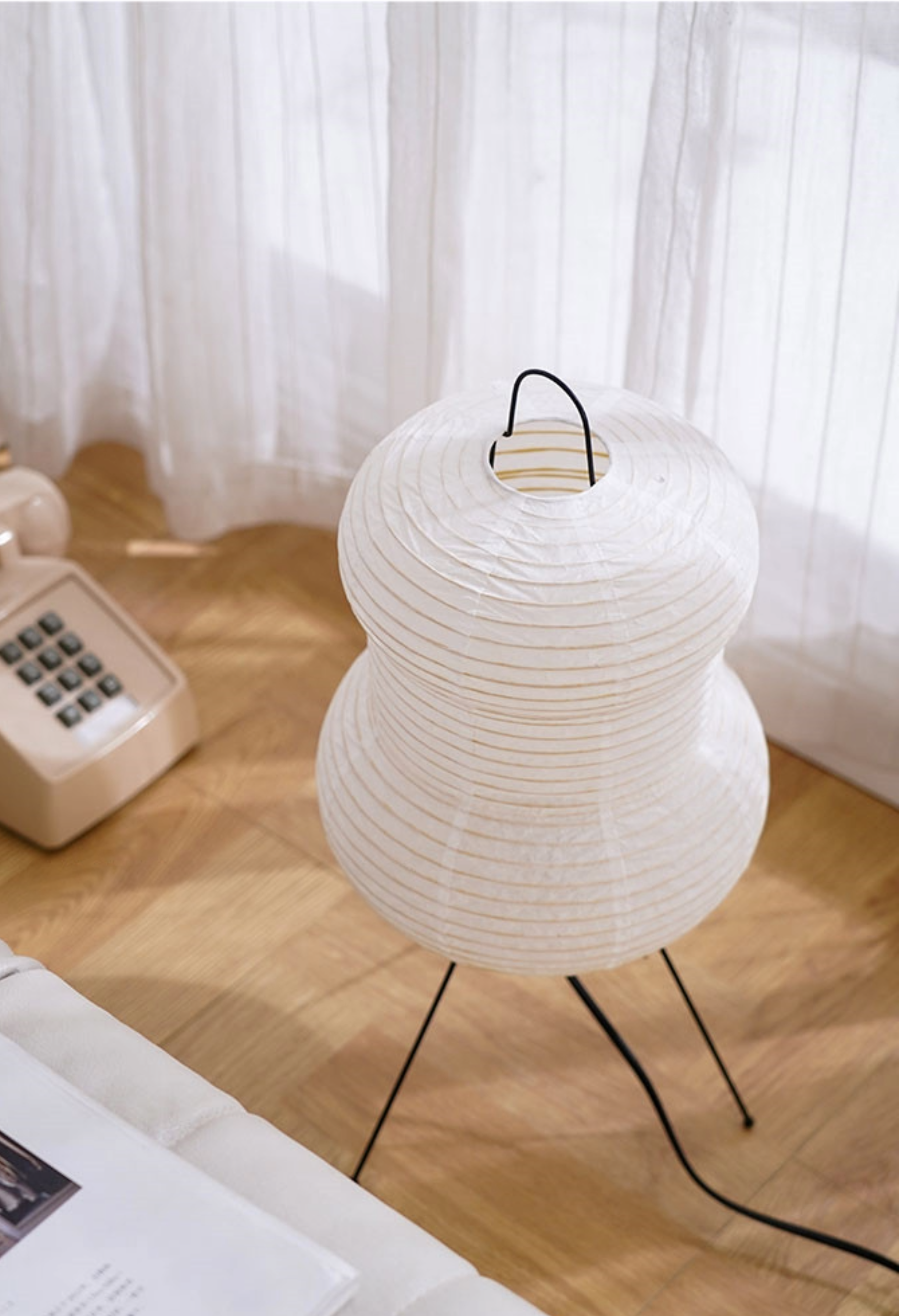 White paper floor lamp with black metal legs on a wooden floor, near white curtains.