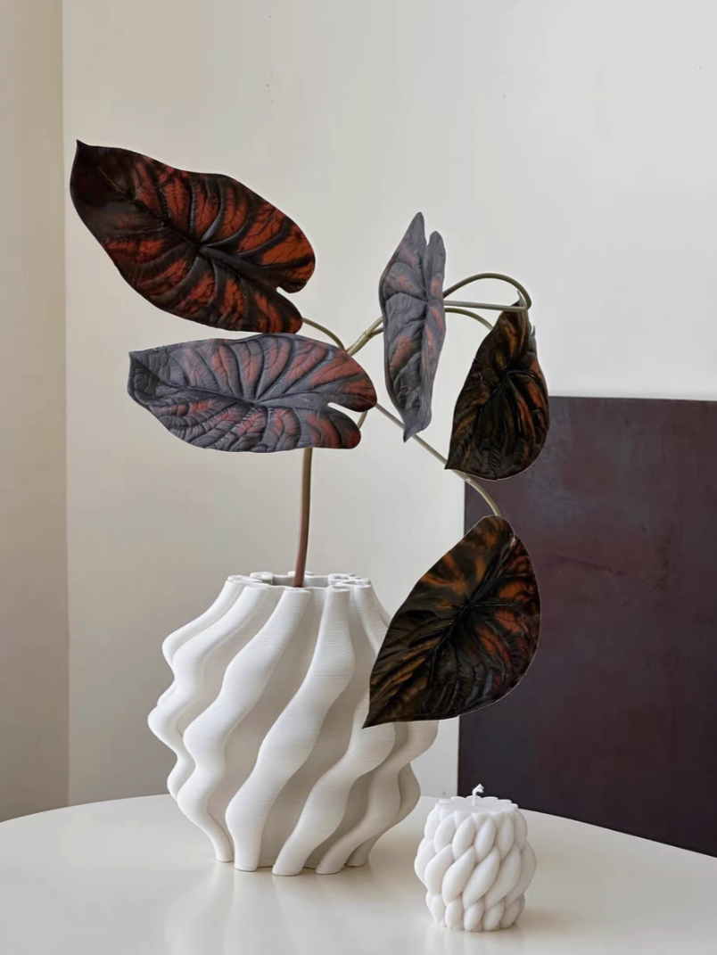 White 3D printed vase with textured design holding dark red-veined foliage.