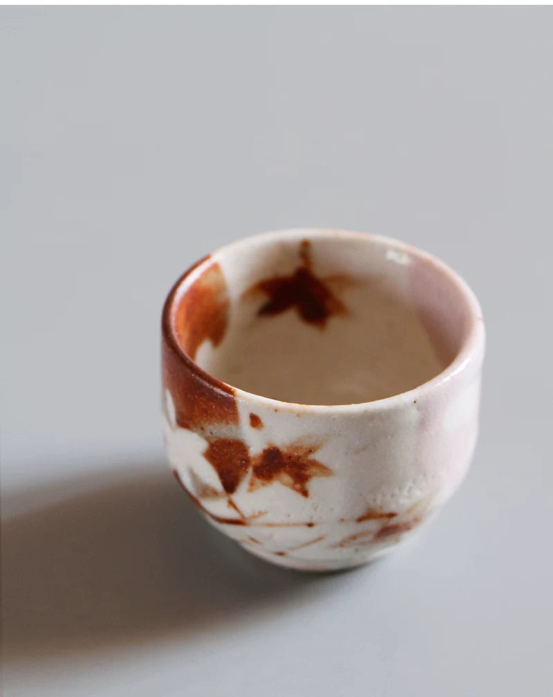 Close-up of a small, round tea cup with a light pinkish interior and reddish-brown maple leaf designs.